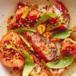 A vibrant dish of grilled meat slices topped with herbs, sliced green onions, red chili flakes, and garlic. The colors are warm, conveying a spicy and savory appeal.