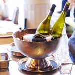A silver ice bucket contains two chilled wine bottles and a rolled cloth napkin on a table, suggesting a relaxed, elegant dining atmosphere.