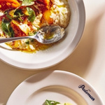 A plate of vibrant risotto topped with roasted vegetables and greens, set on a light background, accompanied by a smaller salad dish with leafy greens.