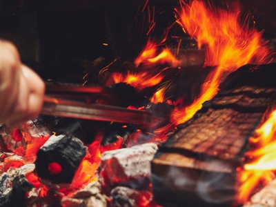 Close-up of hands using tongs to handle glowing charcoal embers and flames on a grill.