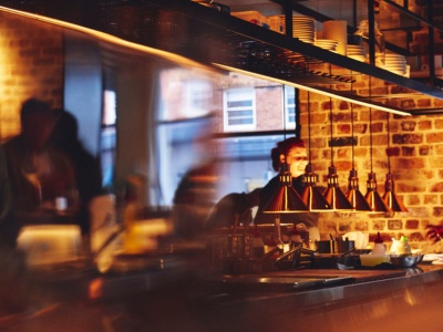 A pair of hands grills meat over a glowing charcoal fire. The scene is rustic and warm, with bright flames and smoke hinting at an outdoor barbecue.