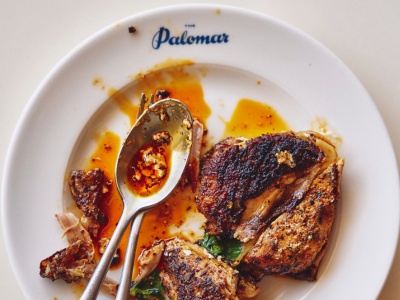 A plate with partially eaten roasted chicken, charred skin, herbs, and a spoon with oil and spices. The Palomar logo is visible, suggesting a restaurant setting.