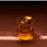 A glass of whiskey with ice and an orange peel garnish sits on a rich brown surface. Warm lighting casts a soft glow, creating an elegant, cozy atmosphere.