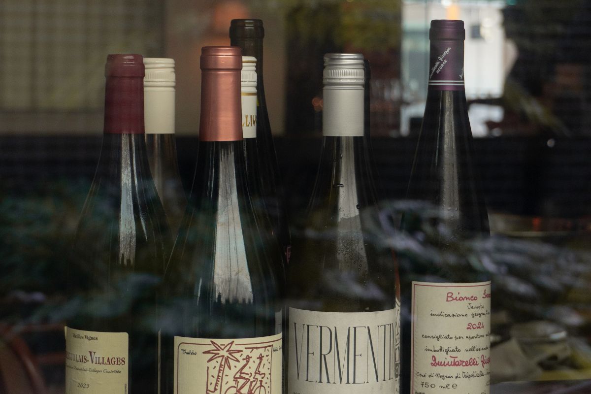 A display of various wine bottles seen through a window. Labels and caps vary in color and design, suggesting a diverse selection. Light reflections add a cozy atmosphere.
