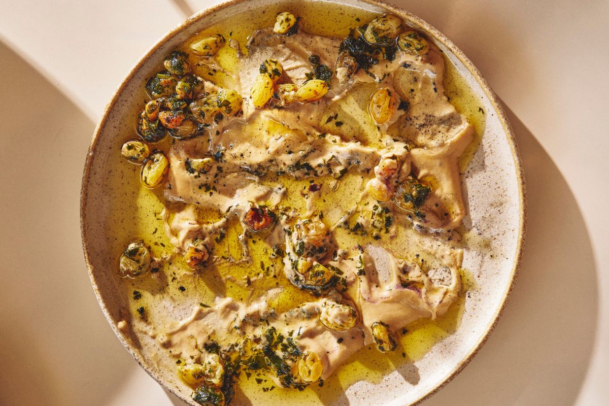 A ceramic plate features creamy fish topped with charred grape halves, herbs, and golden olive oil, creating a rustic and appetizing presentation.