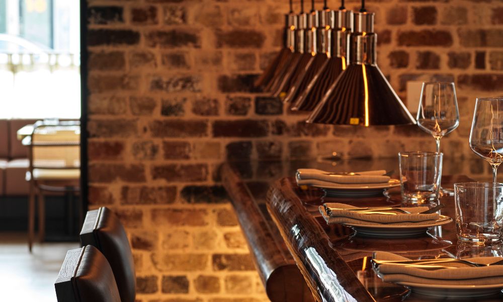 Cozy restaurant scene with a polished wooden bar set for dining, featuring stacked plates, cutlery, and glasses. Warm lighting and exposed brick walls create an inviting atmosphere.