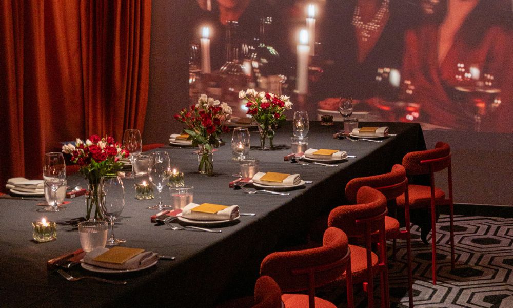 Elegant dining table set for a formal event with red chairs, candles, wine glasses, and flower centerpieces under warm lighting and red curtains.