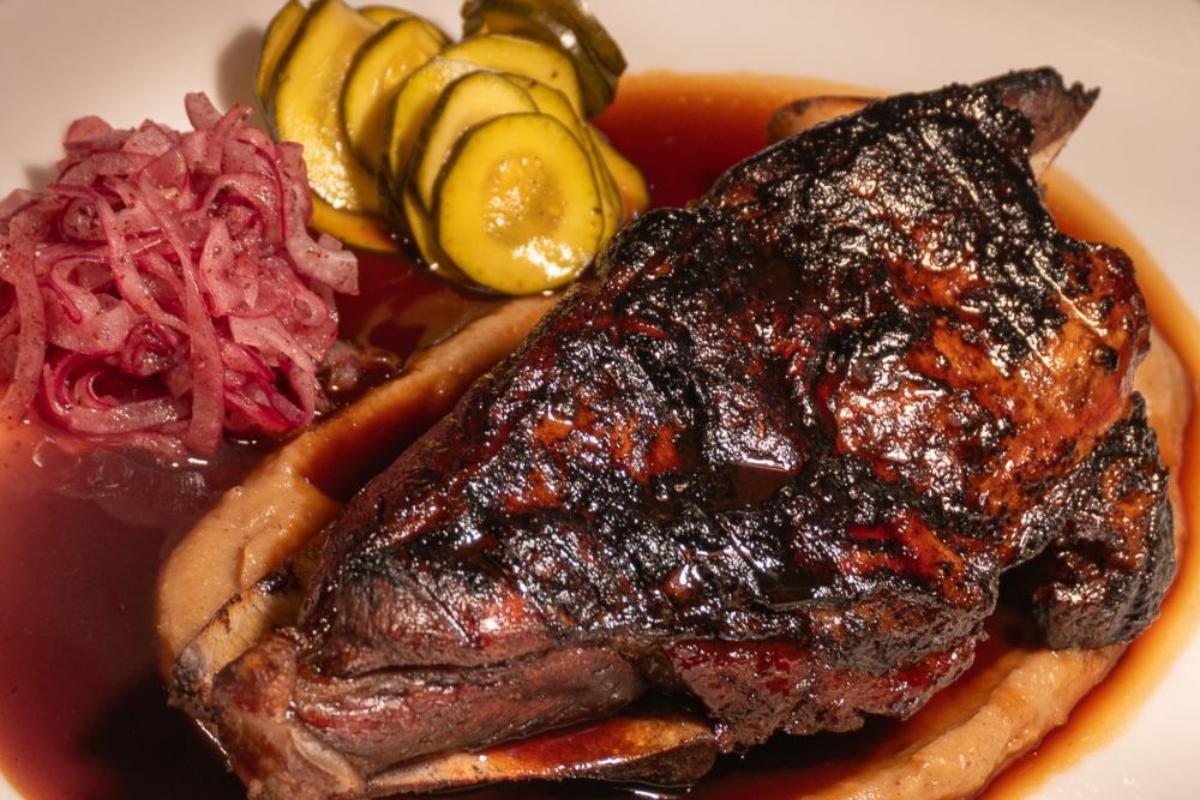 Grilled steak served with sliced pickles, pickled red onions, and a rich brown sauce on a white plate.